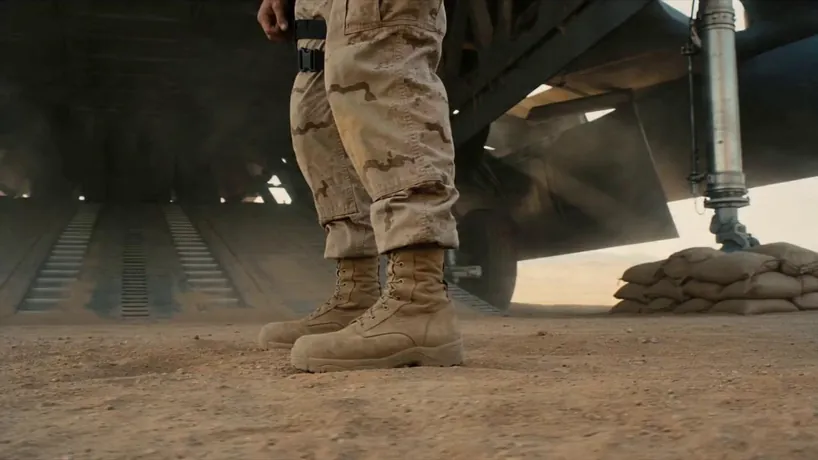 Cinematic VFX sci-fi transformation shot on Arri Alexa Mini LF with a low-angle steady-cam orbit. The scene features a rugged American soldier standing in a dusty desert hangar; as he activates a wrist-mounted neural trigger, heavy matte-black industrial armor plates and hydraulic pistons surge upward from his boots, encasing his body in a militaristic mecha frame. The camera circles closely as copper wires and steel framework interlock with violent precision, capturing his grimace of physical strain. The process accelerates with high-pressure steam venting from shoulder joints as a bulky chest plate slams shut. The transformation concludes with a heavy metallic resonance as a reinforced tactical helmet locks into place. The atmosphere is gritty and visceral, driven by a rhythmic crescendo of clanking metal and pressurized air, ending in a powerful, grounded stance.