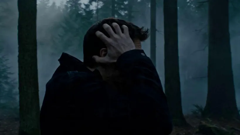 Cinematic dark fantasy VFX transformation shot on Arri Alexa 35 with Master Prime lenses, featuring a continuous high-speed 360-degree orbital camera move around a distressed 30-year-old American man in a dimly lit, foggy forest. The sequence begins with the man clutching his head in agony as his skin ripples and coarse brown fur erupts from his cheeks; his jaw audibly cracks and elongates into a lupine snout while his teeth sharpen into lethal fangs. As the camera revolves, his eyes shift from blue to a glowing predatory amber, and his fingernails transform into jagged black claws. The shot concludes with the fully transformed werewolf letting out a visceral roar toward the full moon as the camera completes its rotation. The atmosphere is dark, claustrophobic, and bone-crunching, with an intense rhythmic pace that mirrors the violent physical evolution.
