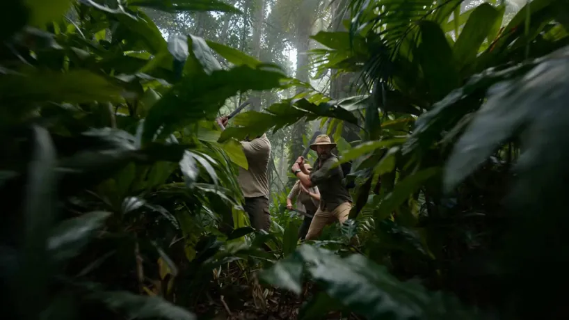 Cinematic survival thriller shot on Arri Alexa 35 with handheld gritty realism and high-tension VFX, featuring fast-paced editing and an immersive, humid jungle atmosphere.
S1: Low-angle tracking shot through dense foliage as a diverse expedition team of three hacks through the humid Amazon undergrowth.
S2: Close-up of the lead explorer freezing in terror as a massive, muddy ripple moves through the dark river water nearby.
S3: Wide handheld shot of a colossal CGI python lunging from the shadows, its scales glistening as it snaps a thick tree branch.
S4: Low-angle whip pan following the serpent's rapid strike toward the camera, mouth agape with razor-sharp teeth.
S5: Extreme close-up of a team member screaming as they dive into the mud, water splashing everywhere in a chaotic struggle.
S6: Medium shot of the python's massive body coiling around abandoned gear, crushing it instantly with a loud, resonant crunch.
S7: High-angle wide shot of the team sprinting into the misty jungle as the predator vanishes back into the emerald canopy.
